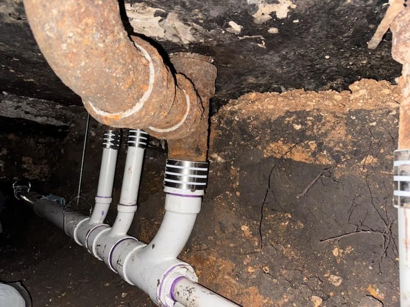 Underground plumbing repair: rusty pipe connected to new PVC pipes with silver couplings in a dirt crawl space.
