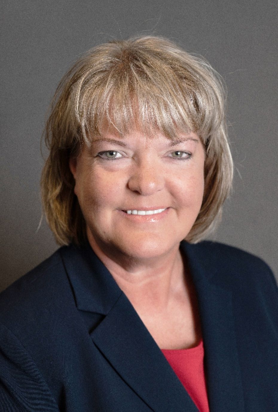 Woman with short, blonde hair smiles, wearing a navy blazer over a red top.