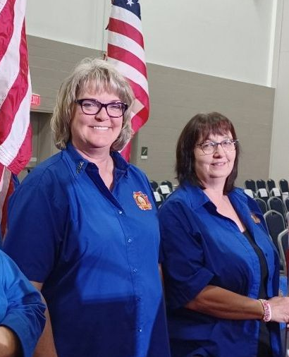 Two women in blue shirts stand before US flags. They smile in a large hall with chairs.