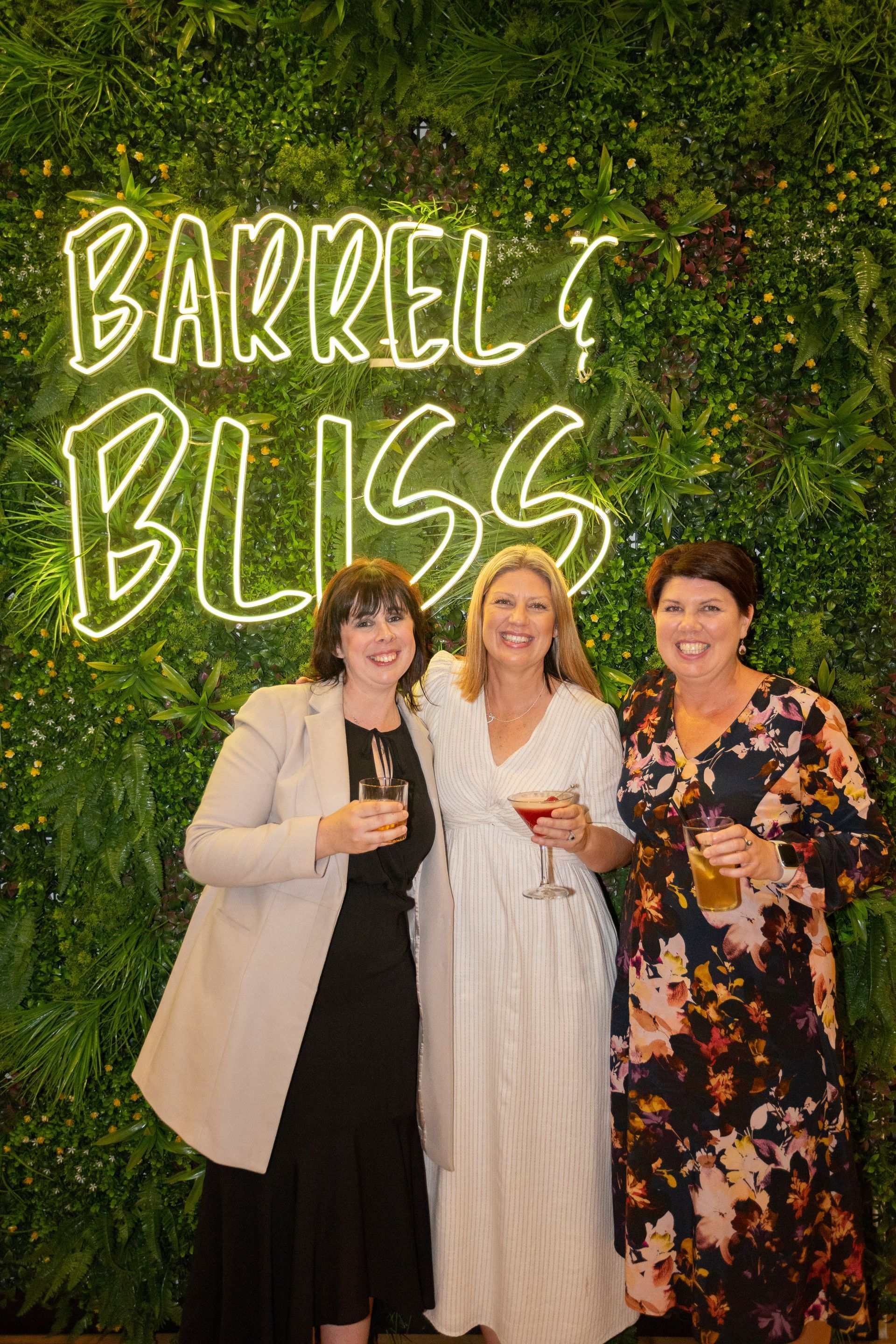 Three women smiling, holding drinks, pose in front of a green wall with neon sign reading 