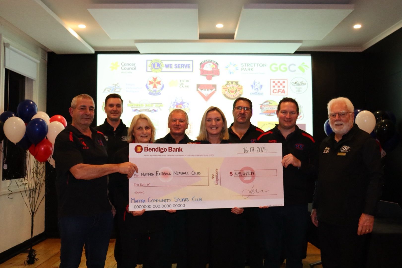 Group of people holding a large check with a backdrop of logos, possibly a donation.