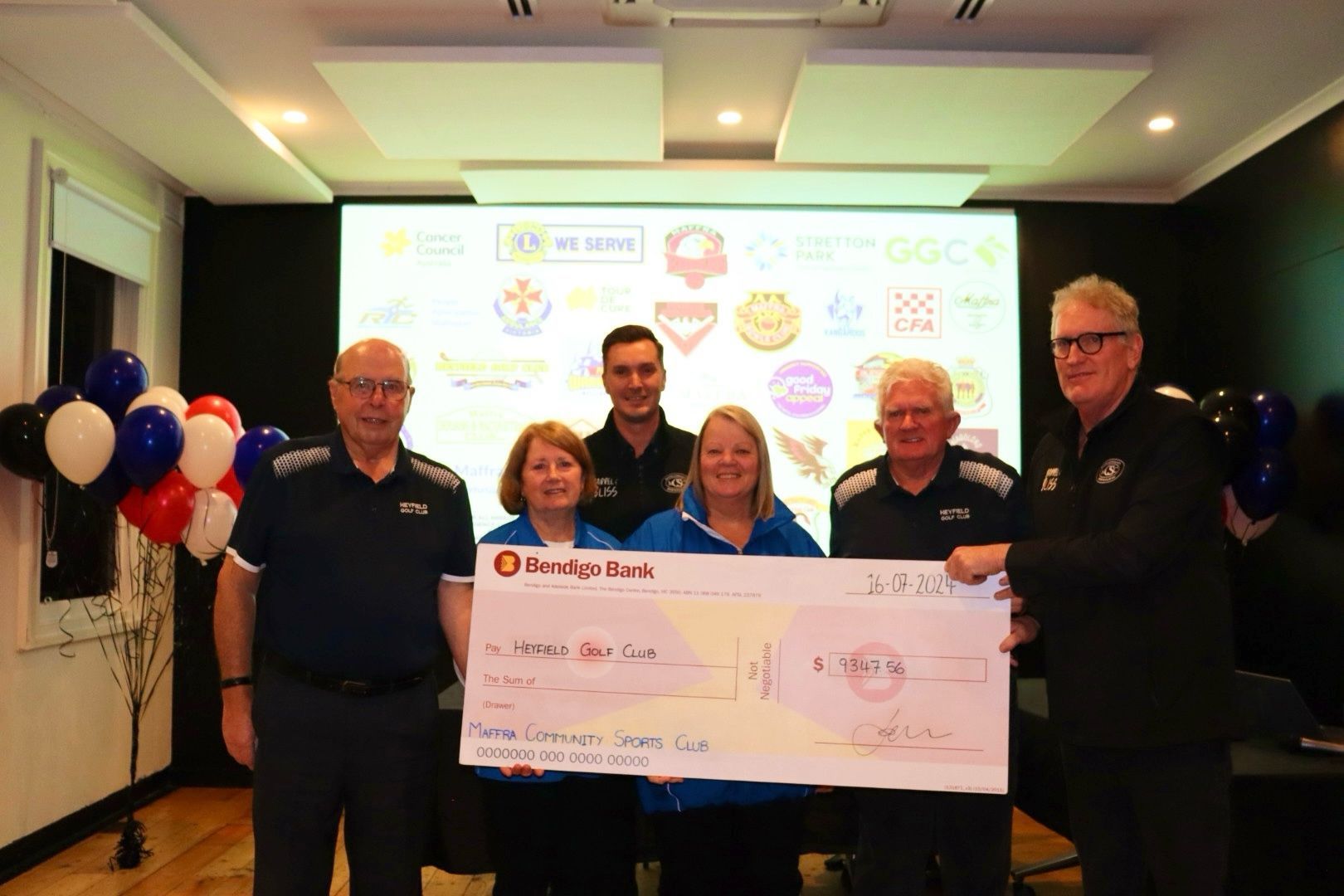 People presenting a large check to a charity, with a logo-filled screen in the background.