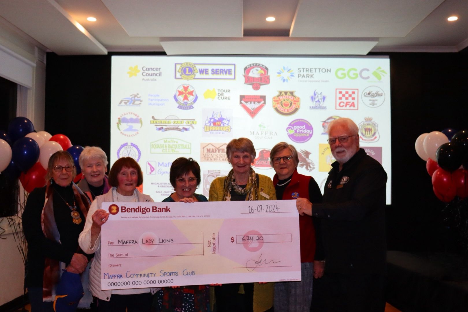 Group holding a large check in front of logos, balloons, and a screen. People smiling.