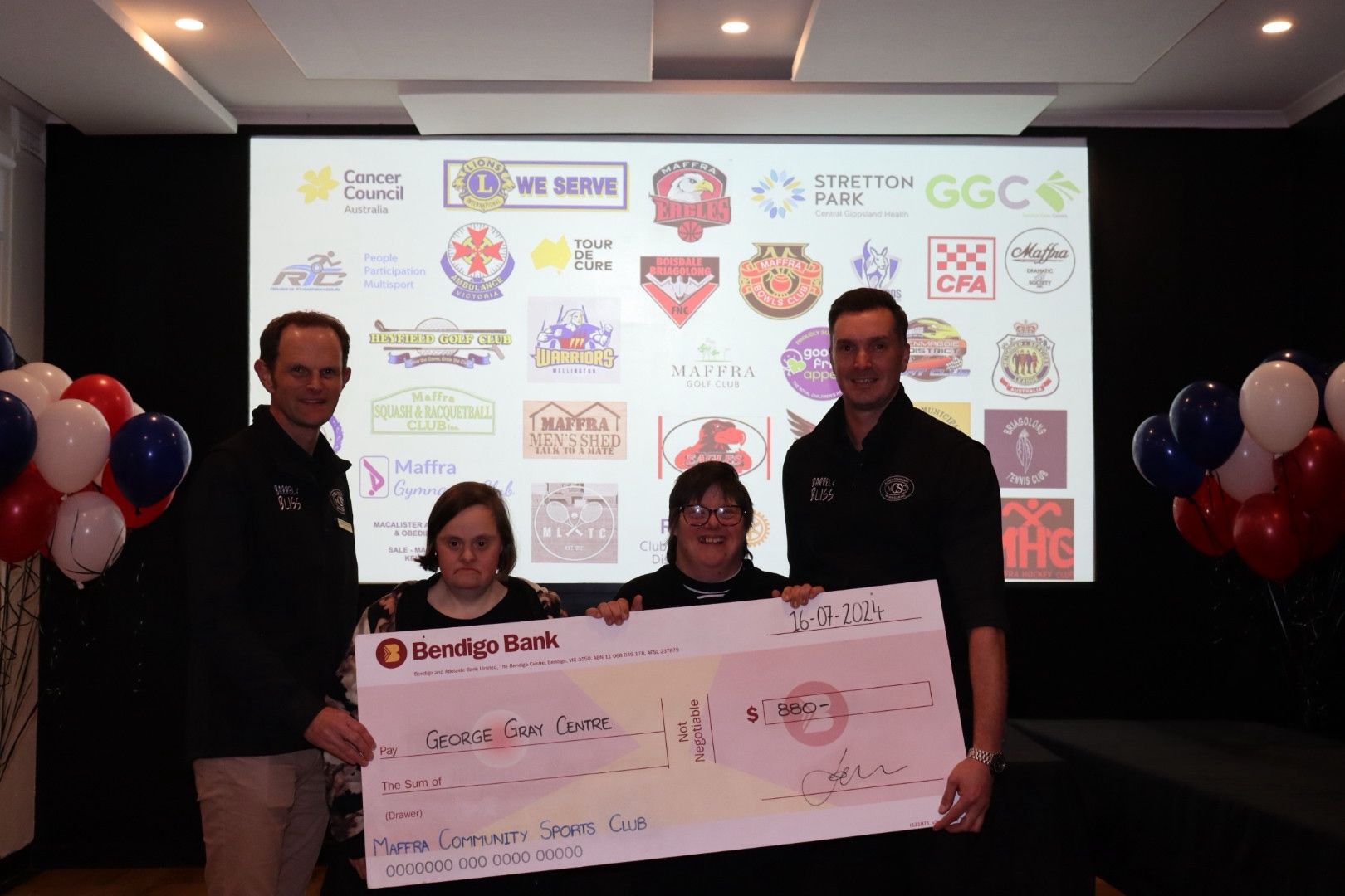 Four people hold a large check in front of a logo-filled screen. Balloons flank them.