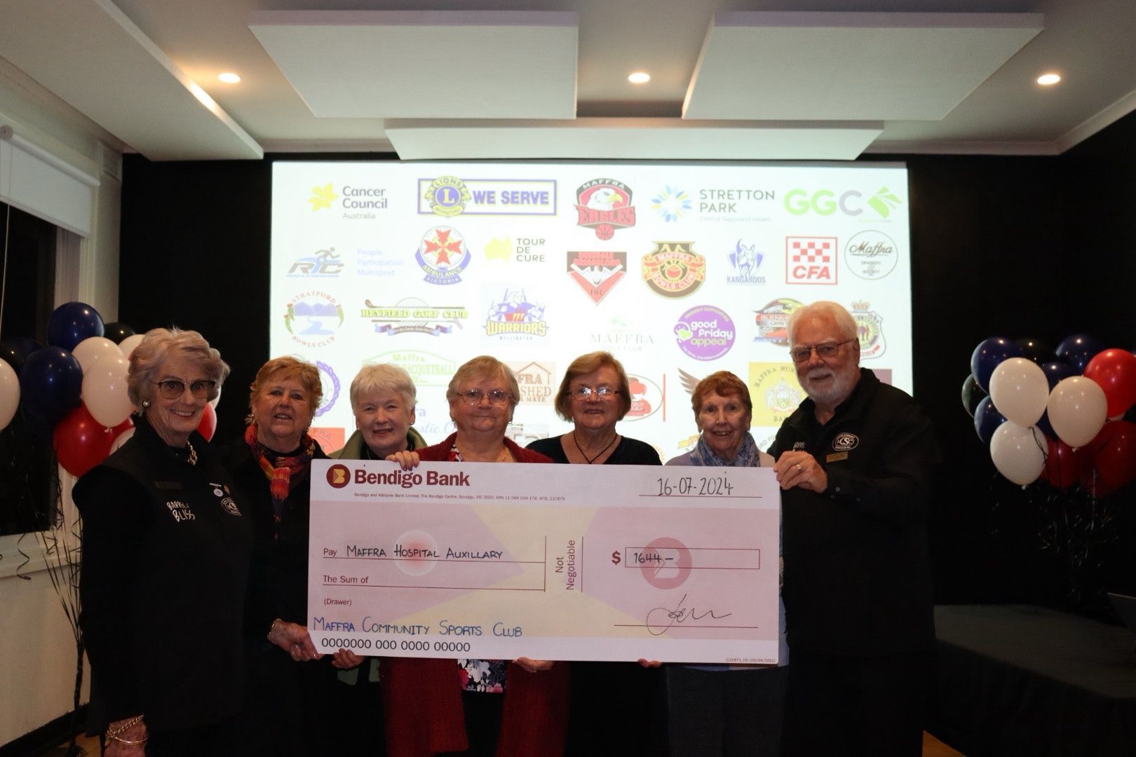 Group presents a large check. They stand in front of a screen with logos. Balloons on either side.