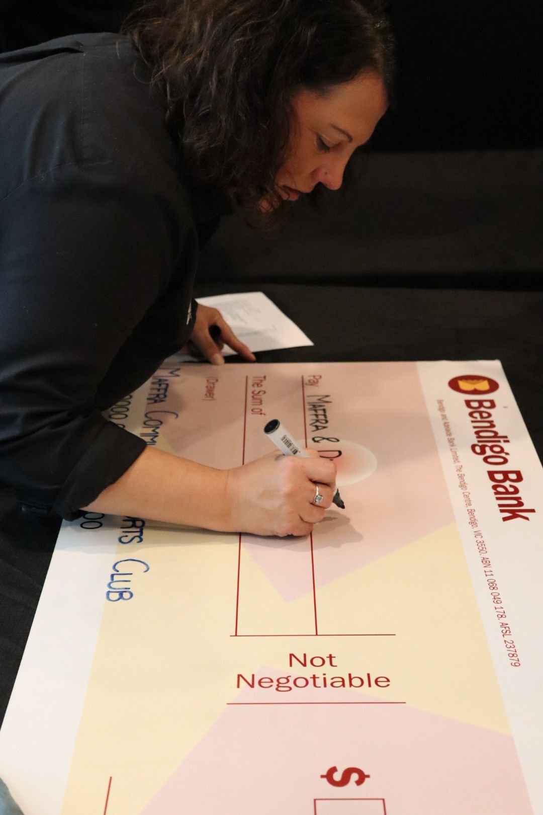 Woman signing a large novelty check from Bendigo Bank, with a pen, indoors.