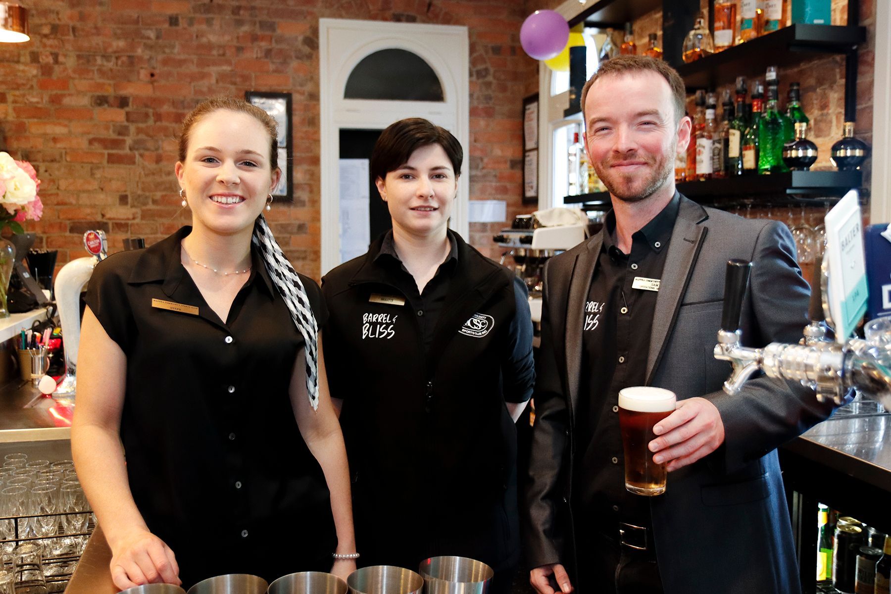 Three people behind a bar. Woman with braided hair and woman with short hair stand on either side of a man holding a beer.