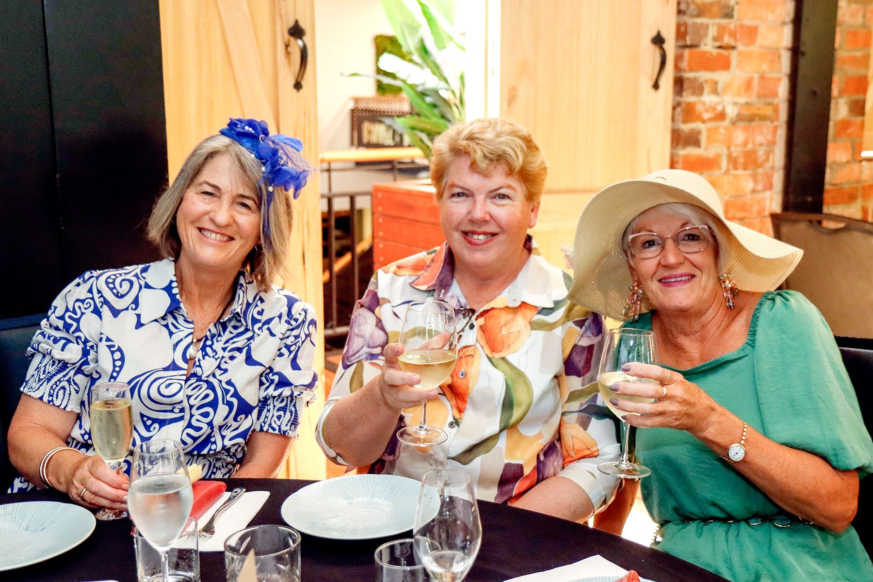 Three women in hats at a table, toasting with drinks. Bright clothing, smiles, restaurant setting.