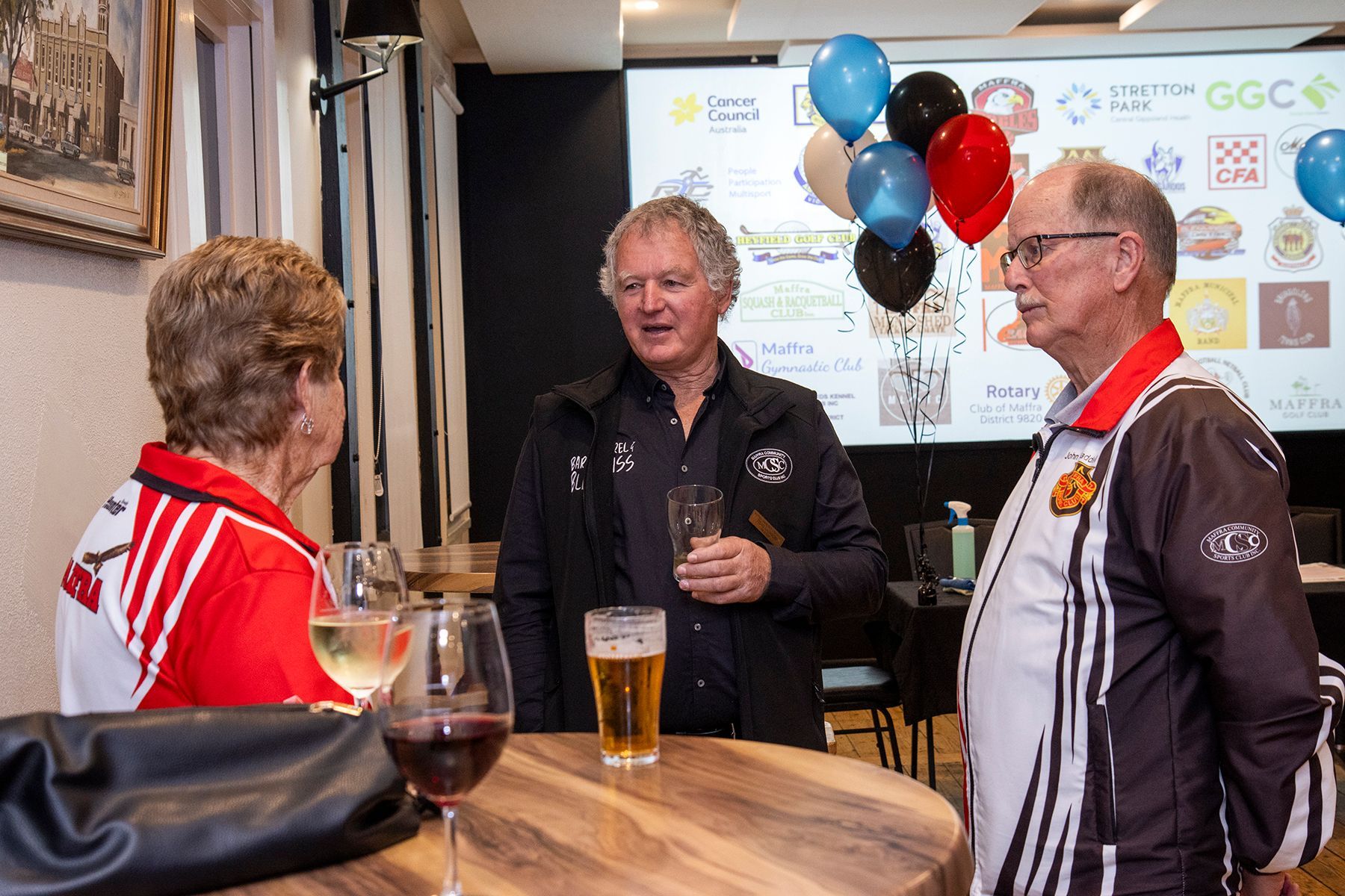 Three people talking, holding drinks, at an event with balloons and sponsor logos.