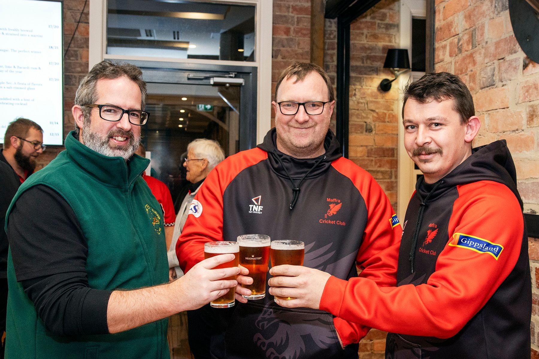 Three men toast with beer in a pub. Two wear red jackets, one a green vest.