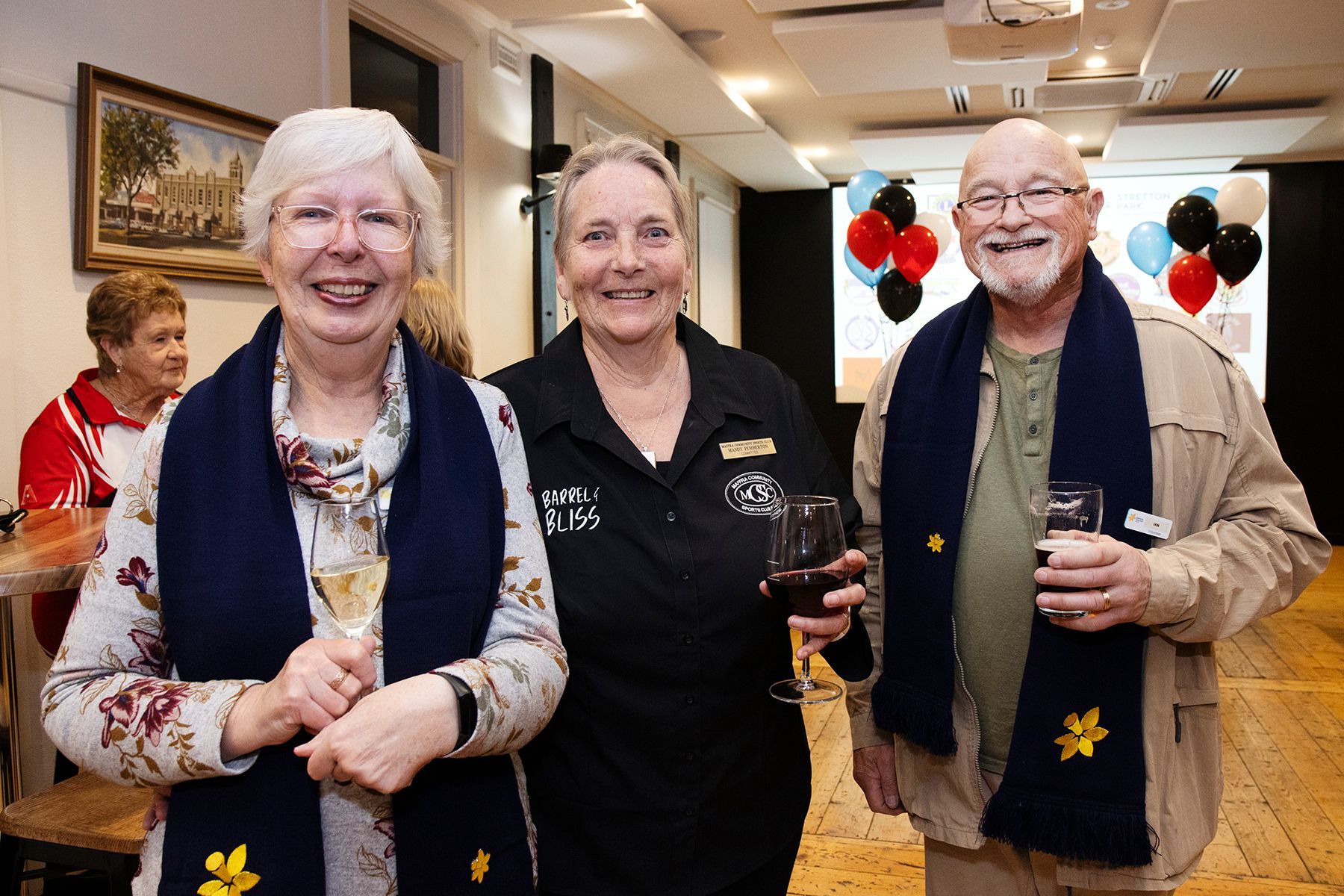 Three people smiling, holding drinks, wearing scarves with flower detail at an event with balloons.