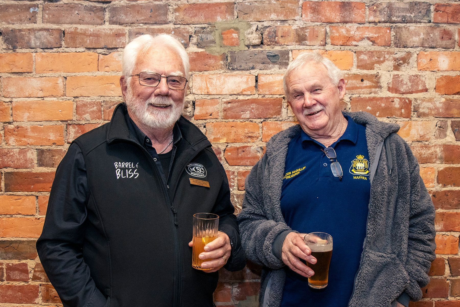 Two older men smiling, holding beer glasses, standing against a brick wall.