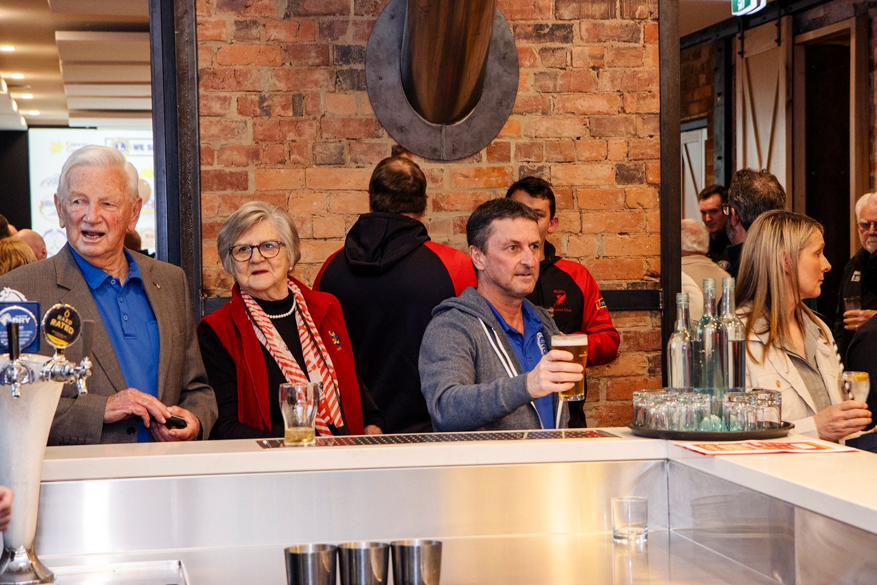 People at a bar, some holding drinks, brick wall background.