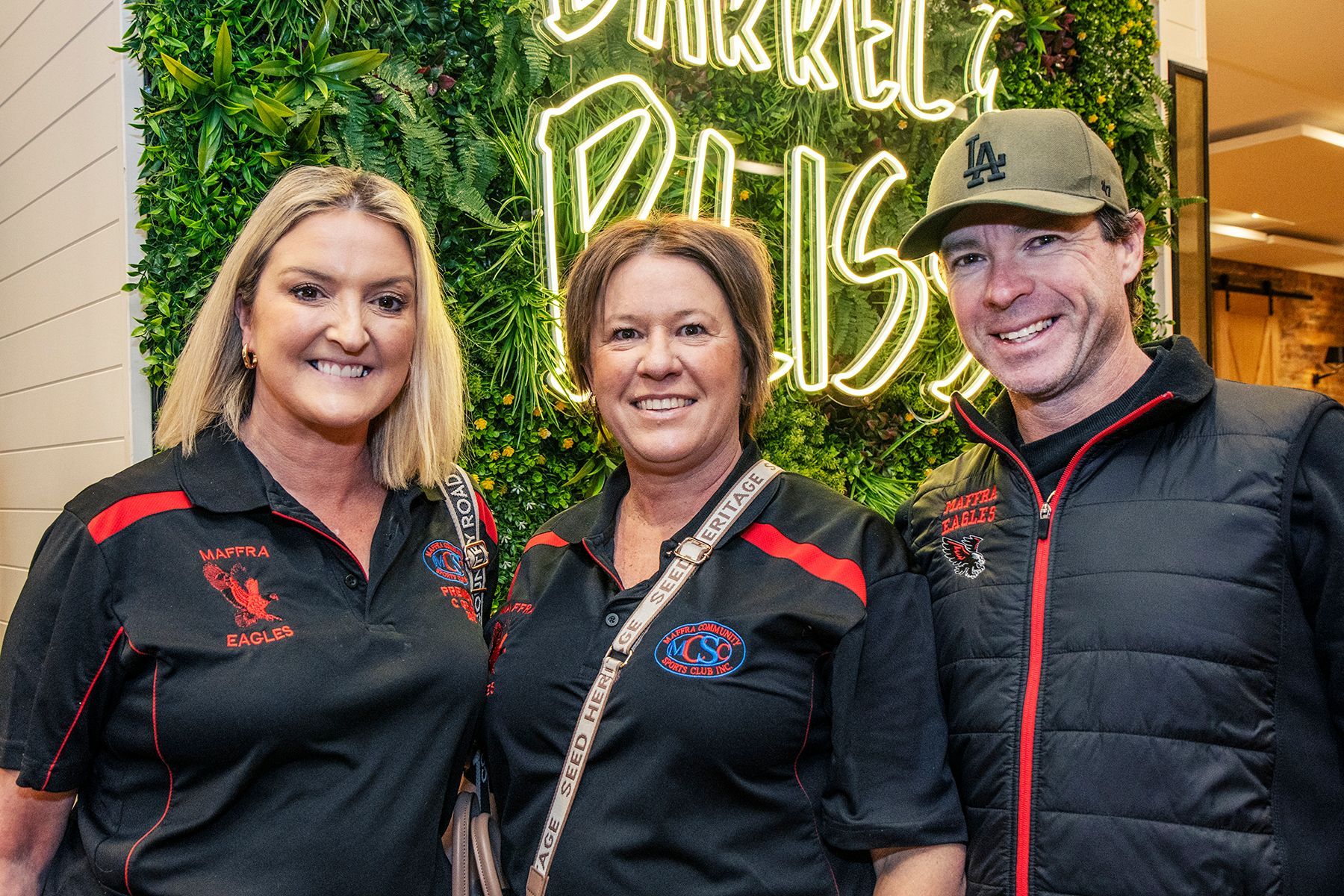 Three people smiling in front of a neon sign at Barrel & Riser. Two women and one man, all wearing black shirts.