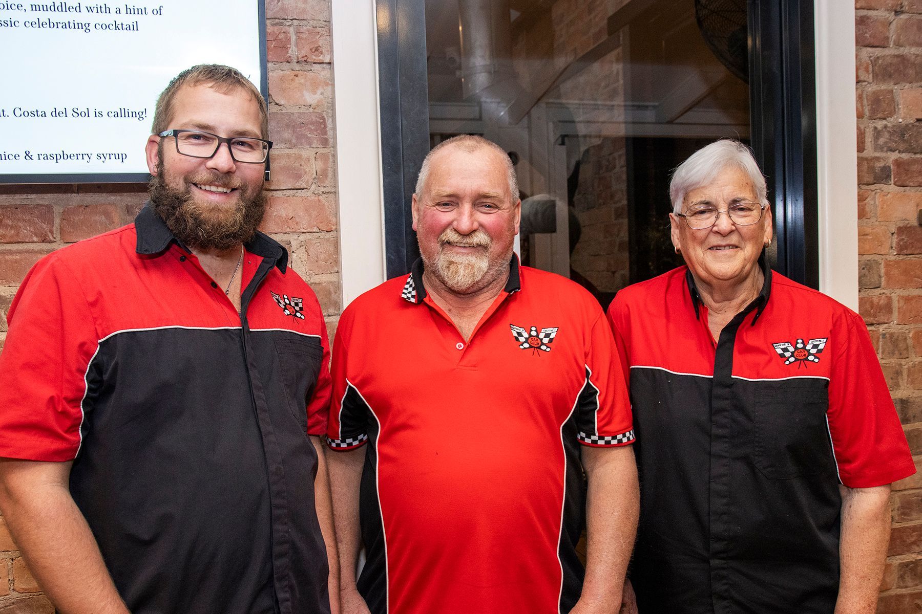 Three people wearing red and black polo shirts, smiling, standing indoors near brick wall.
