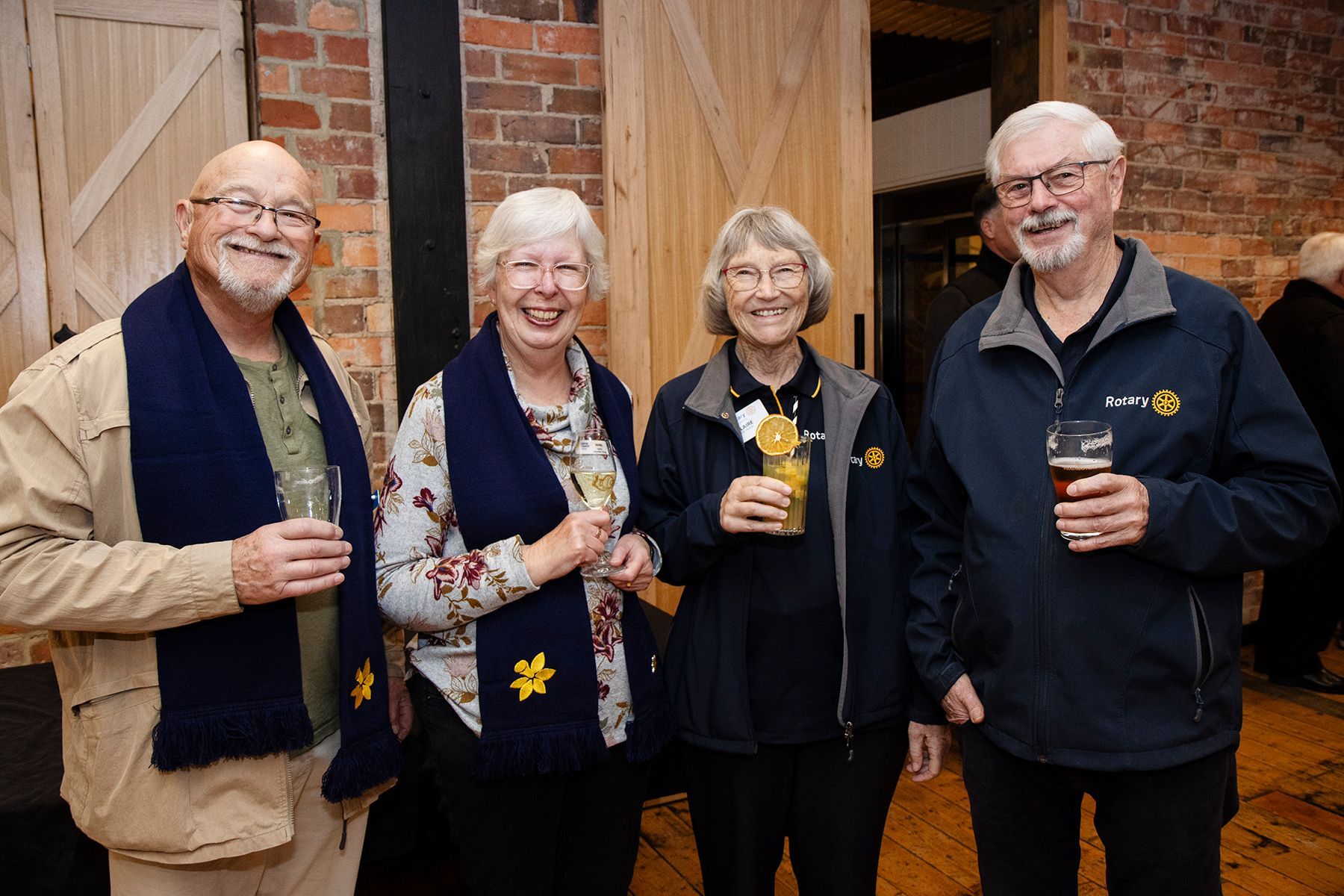 Four smiling people, two men, two women, in a room with brick wall, holding drinks.