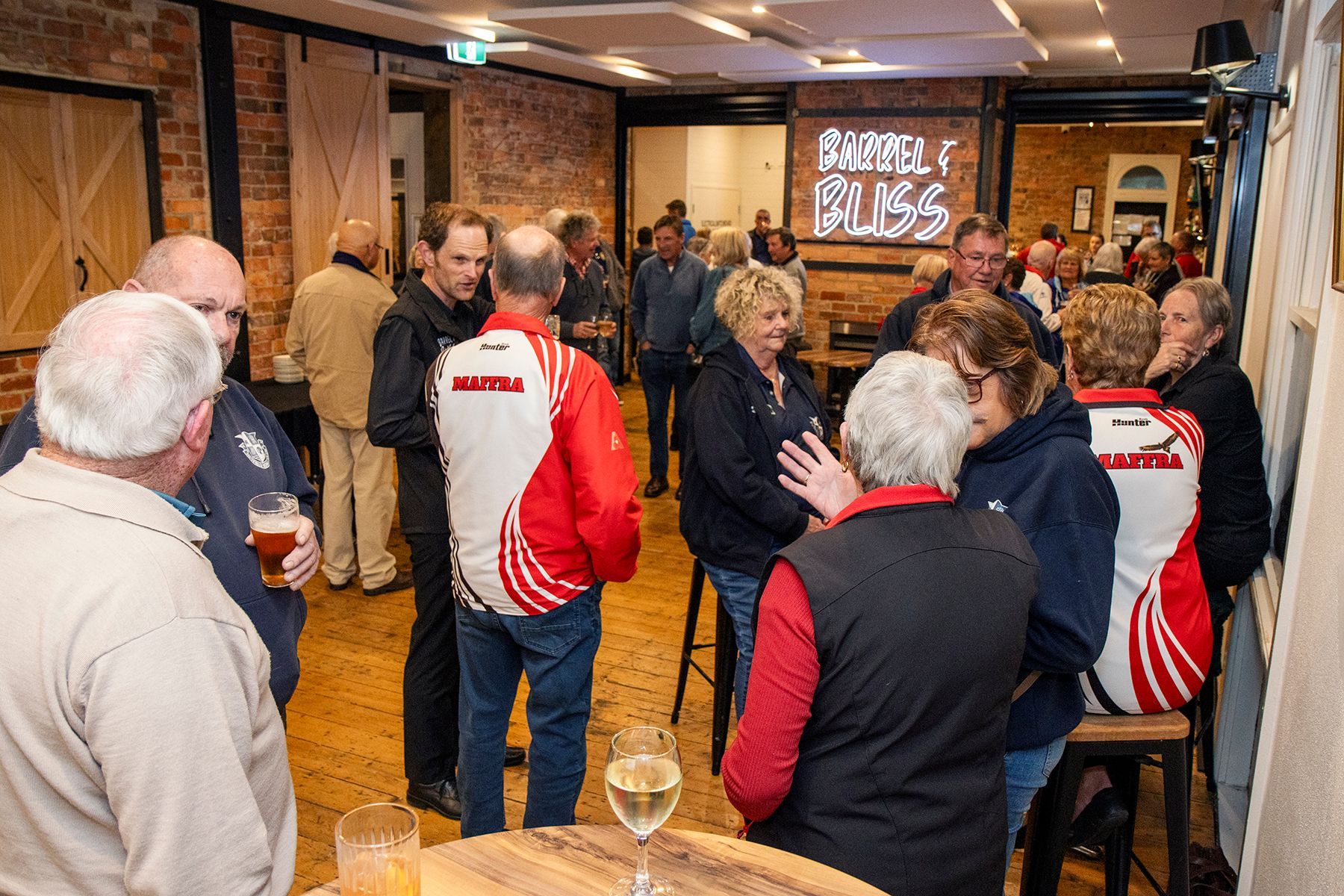 People socializing in a dimly lit room, with drinks. Several wearing matching red and white jackets, near a bar.