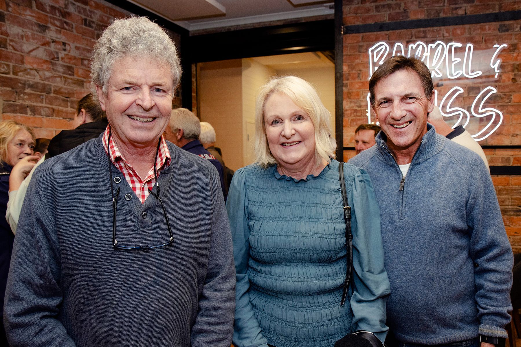 Three smiling people pose in a dimly lit bar. Two men and a woman stand in front of a brick wall with a neon sign.