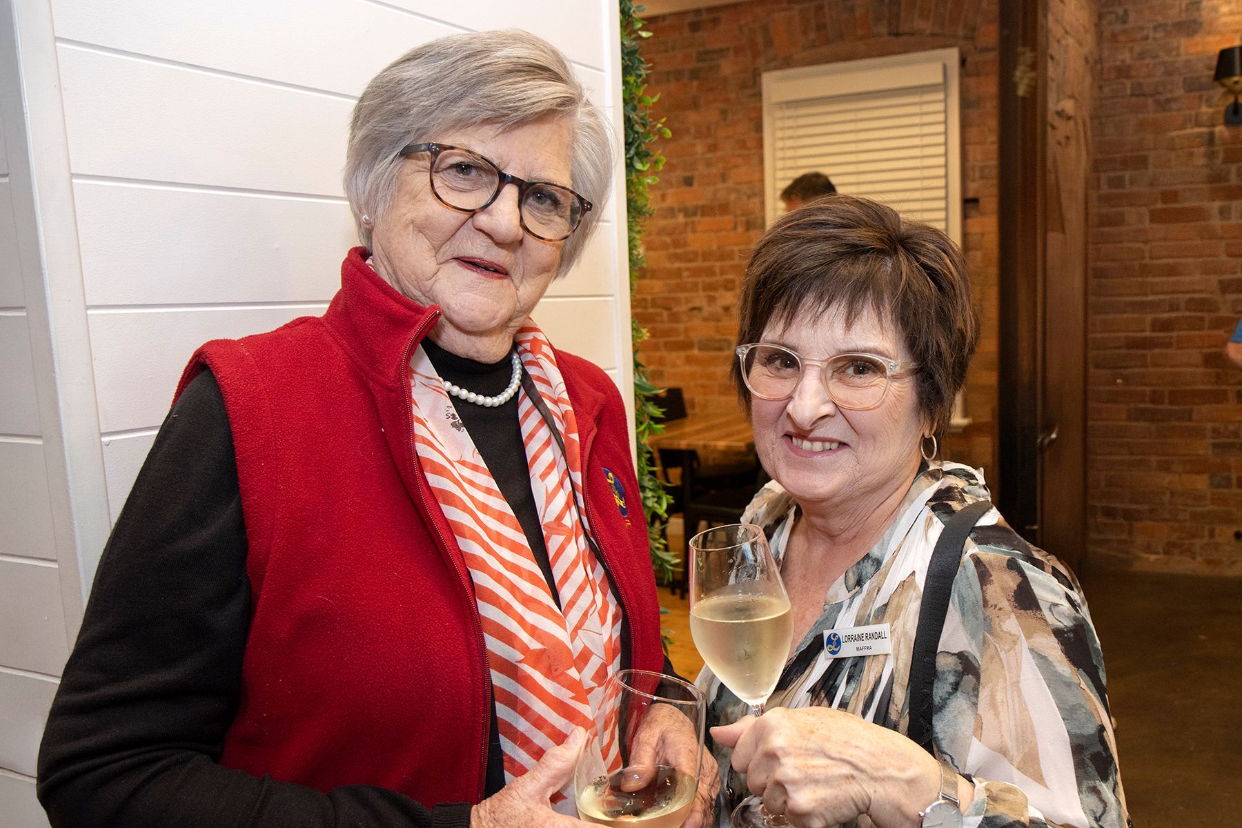 Two women smiling, holding drinks. One in red vest, scarf, glasses. Other in patterned shirt, glasses. Indoors.