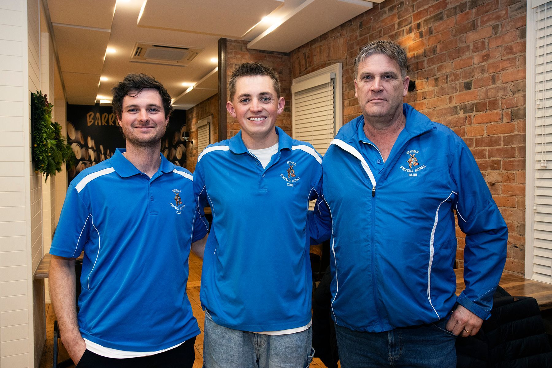Three men in blue shirts standing indoors, smiling, brick wall in background.