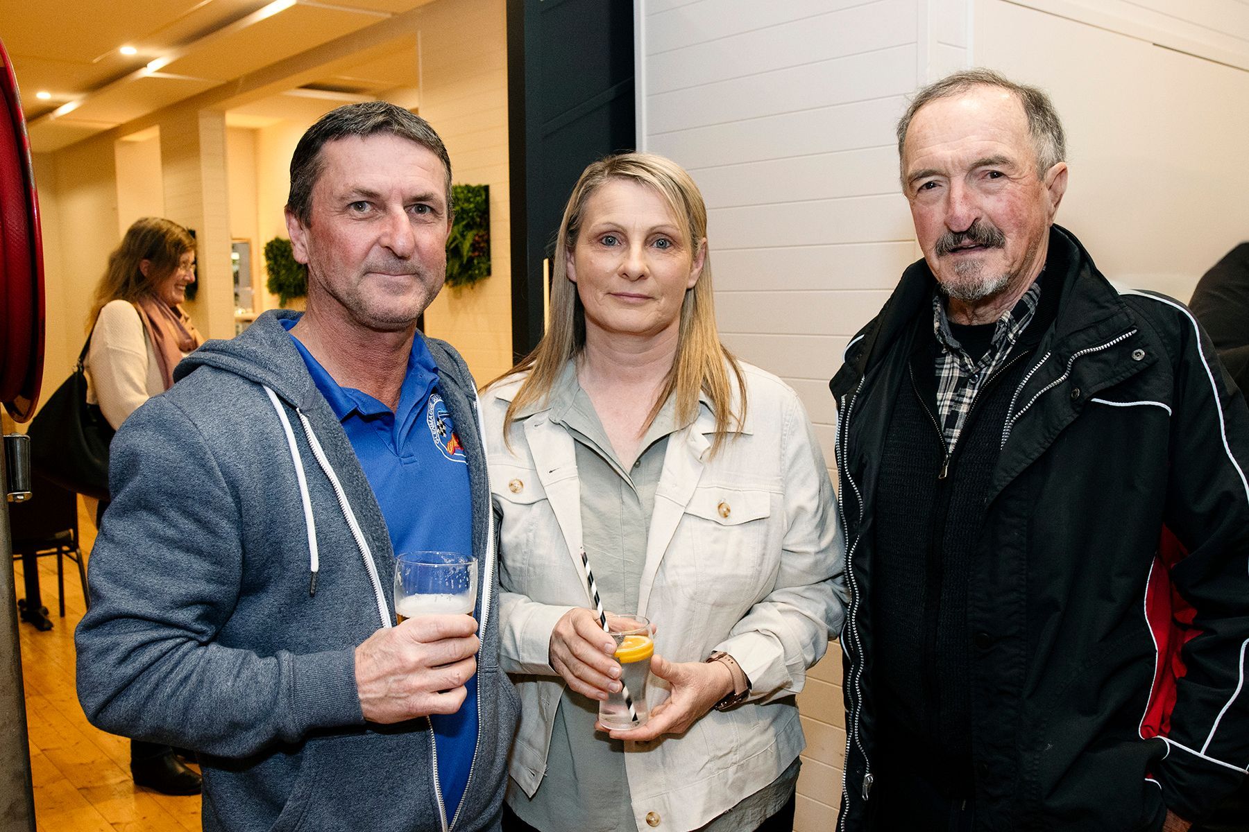 Three people standing together: two men and a woman. The woman in a tan jacket stands between the men, all holding drinks.