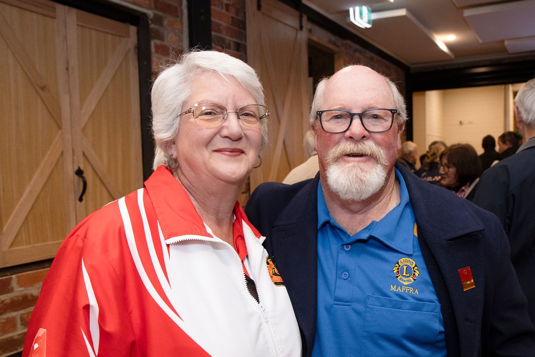 Couple smiling, man with beard, woman in red jacket, posing at event with wood backdrop.