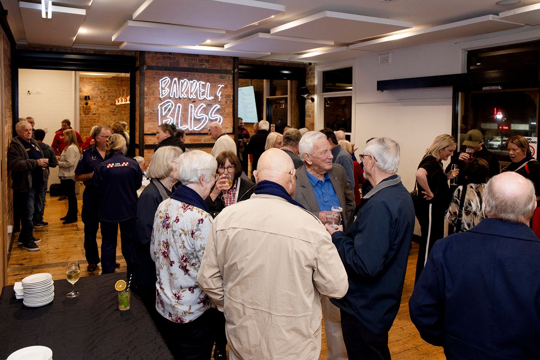 People socializing in a room, some holding drinks. Neon sign on brick wall, dark setting.