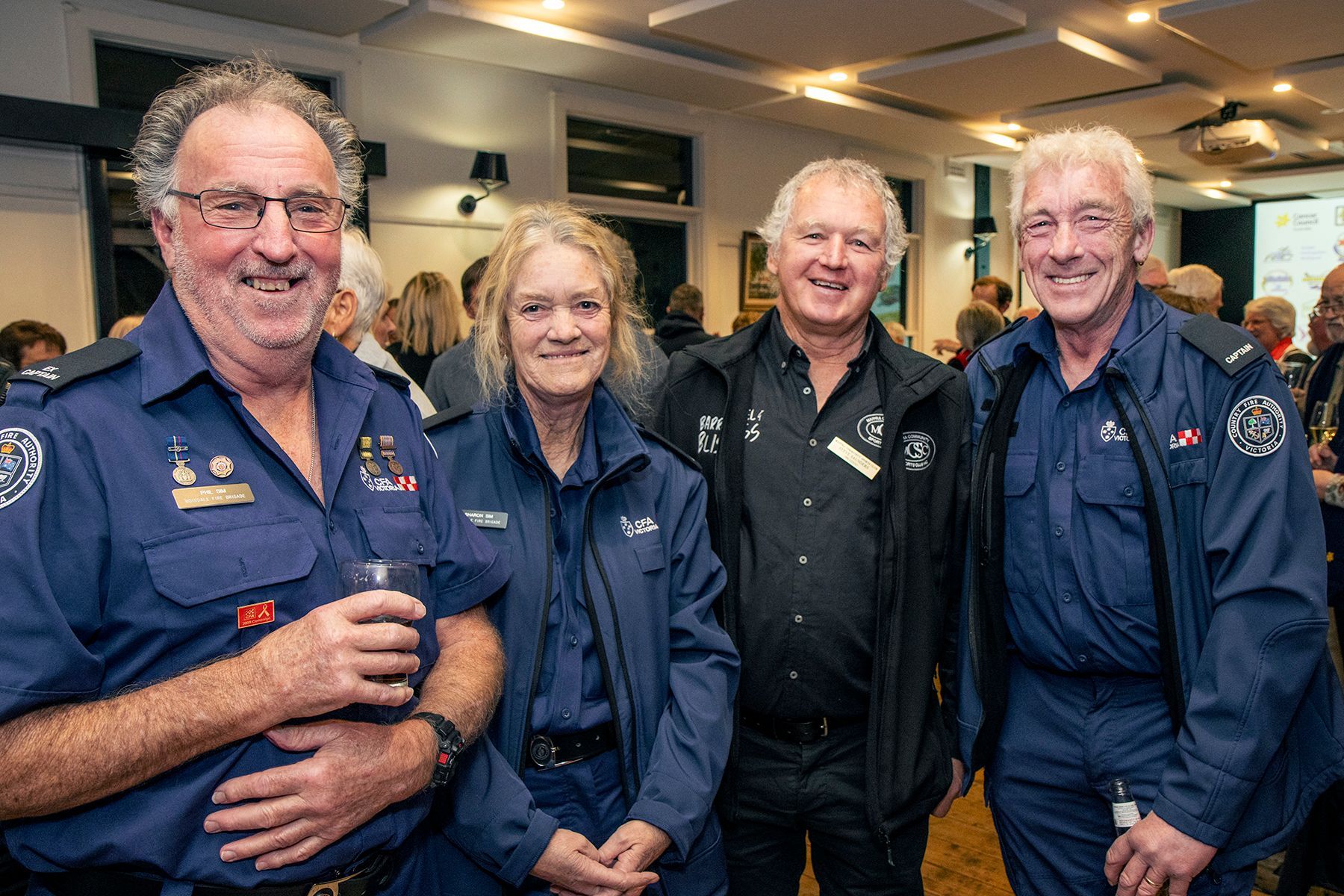 Four people in blue uniforms at an event, smiling. One holds a drink. Background: other attendees.