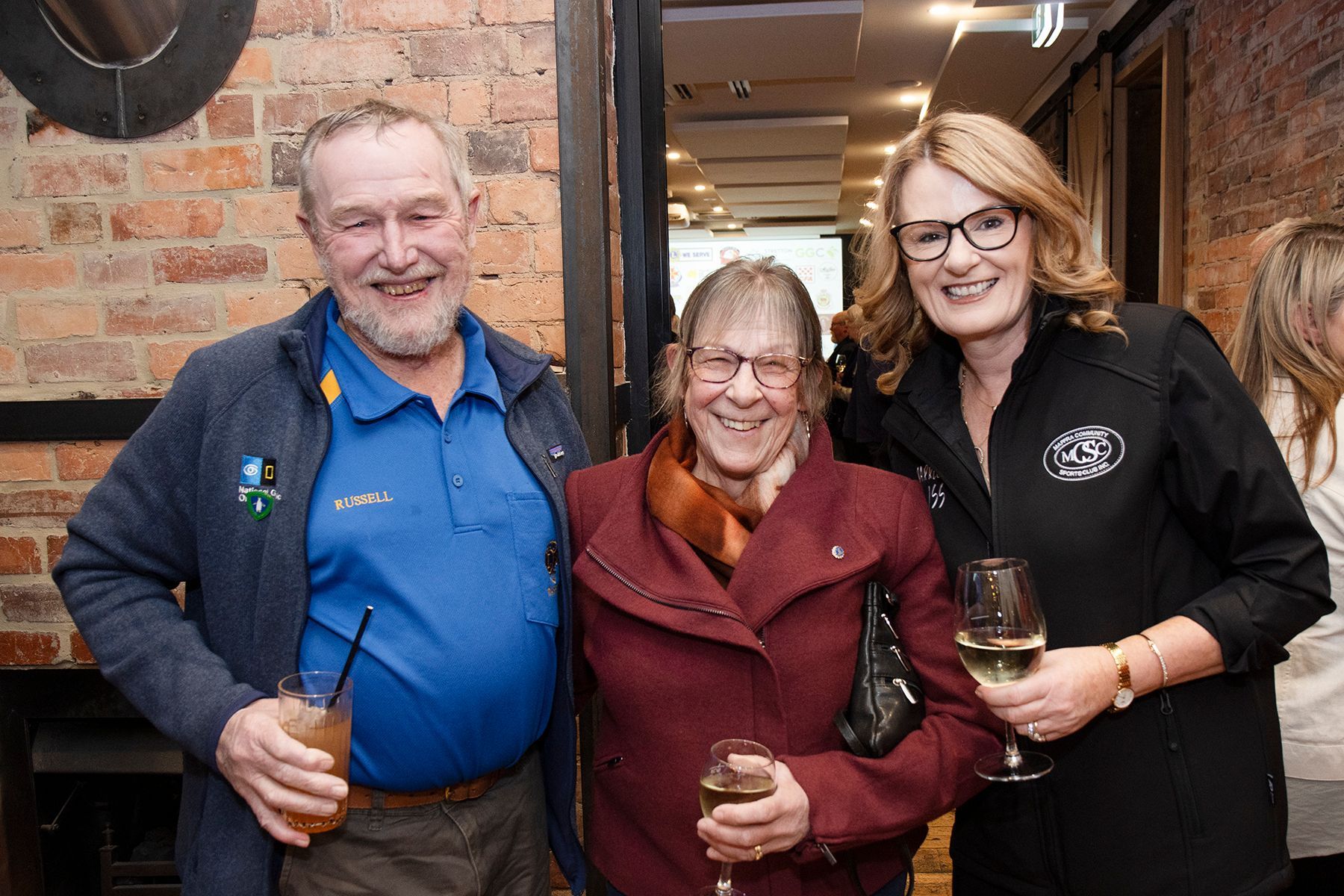 Three people smiling and posing for a photo at an event. Man with blue shirt, woman in red coat, and woman in black jacket.
