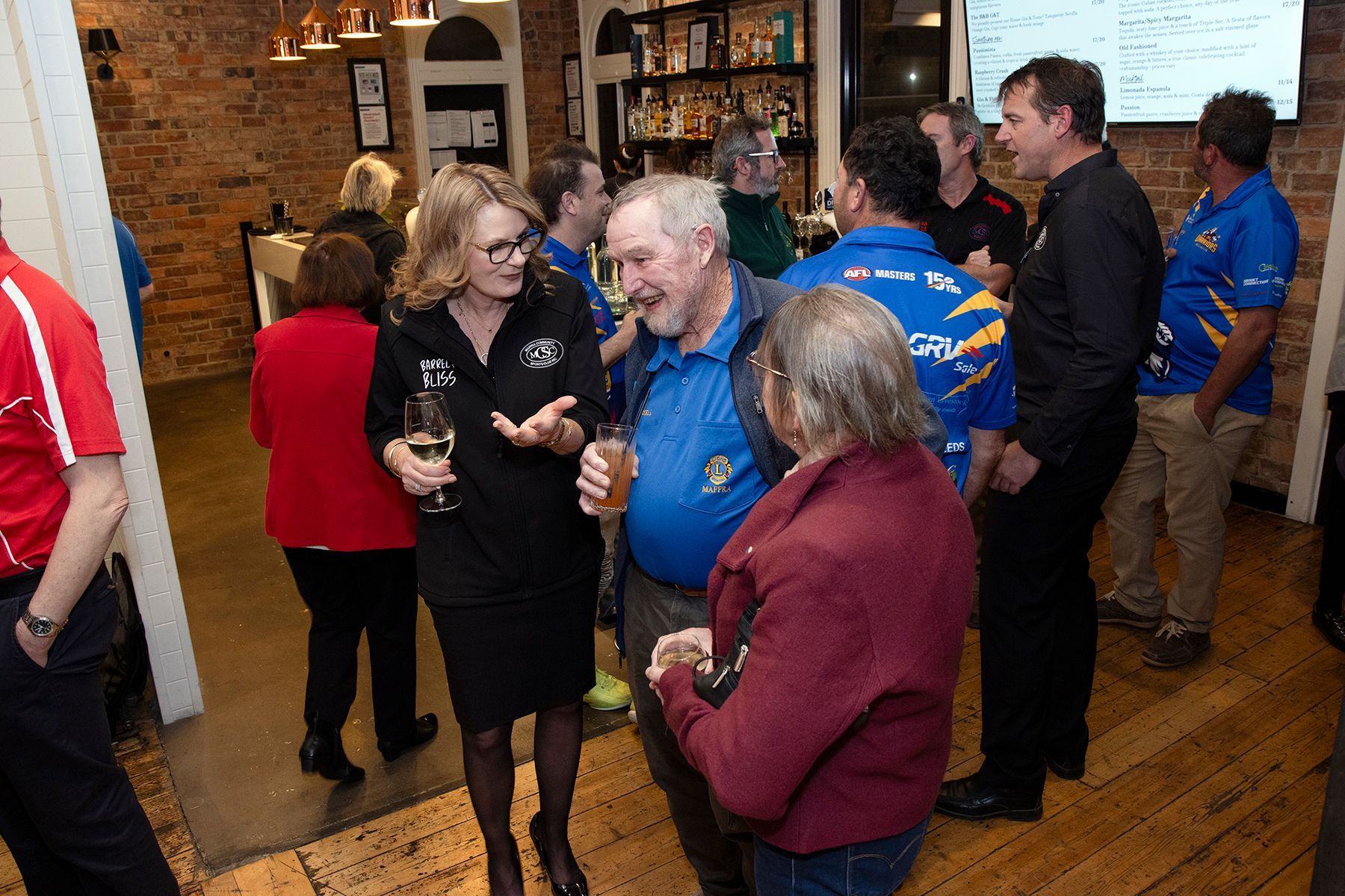 People socializing in a bar. Woman in black speaks to a man; others chat, drinks in hand. Brick walls, wood floor.