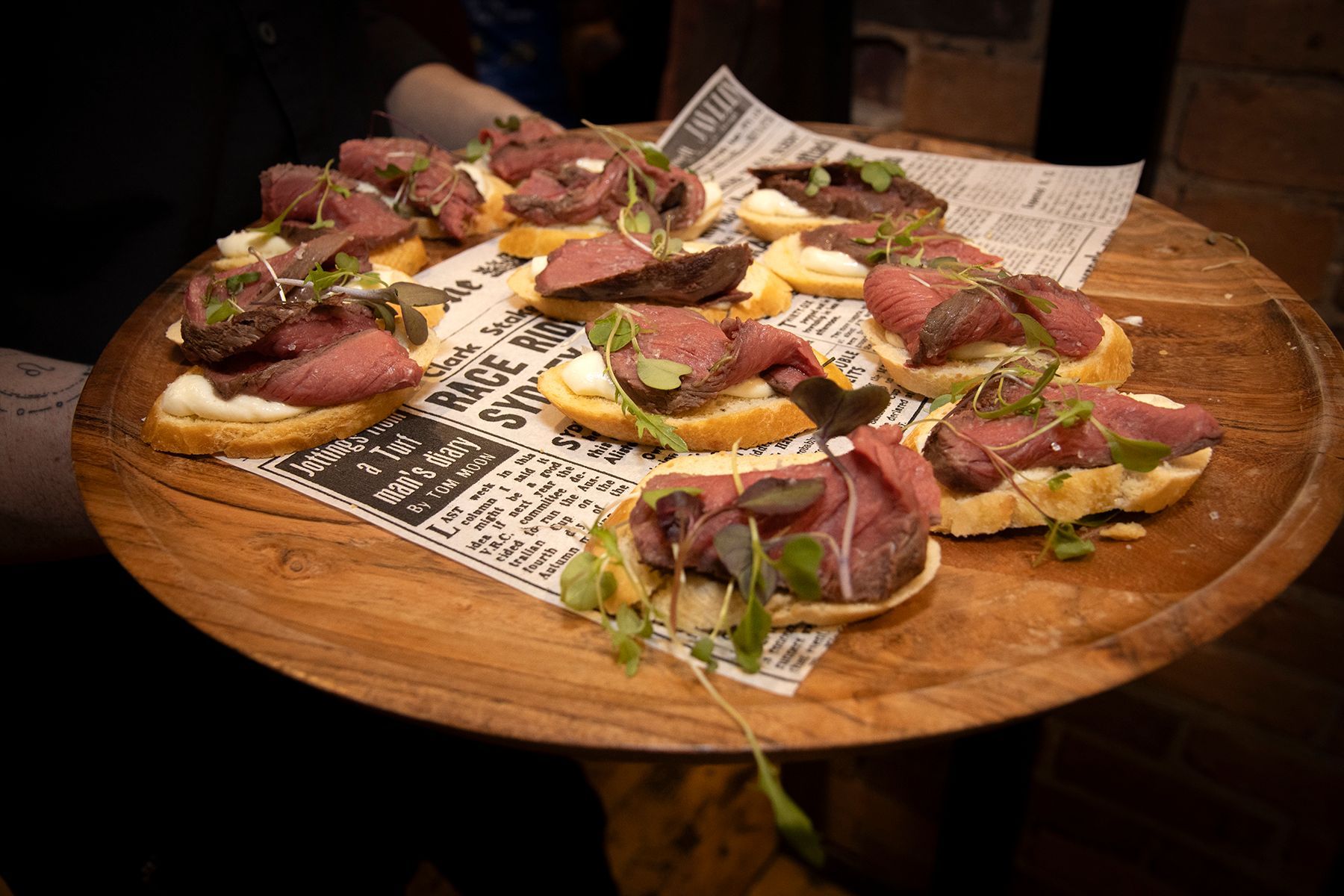 Appetizers on a wooden platter: bread topped with meat, cream cheese, and microgreens.