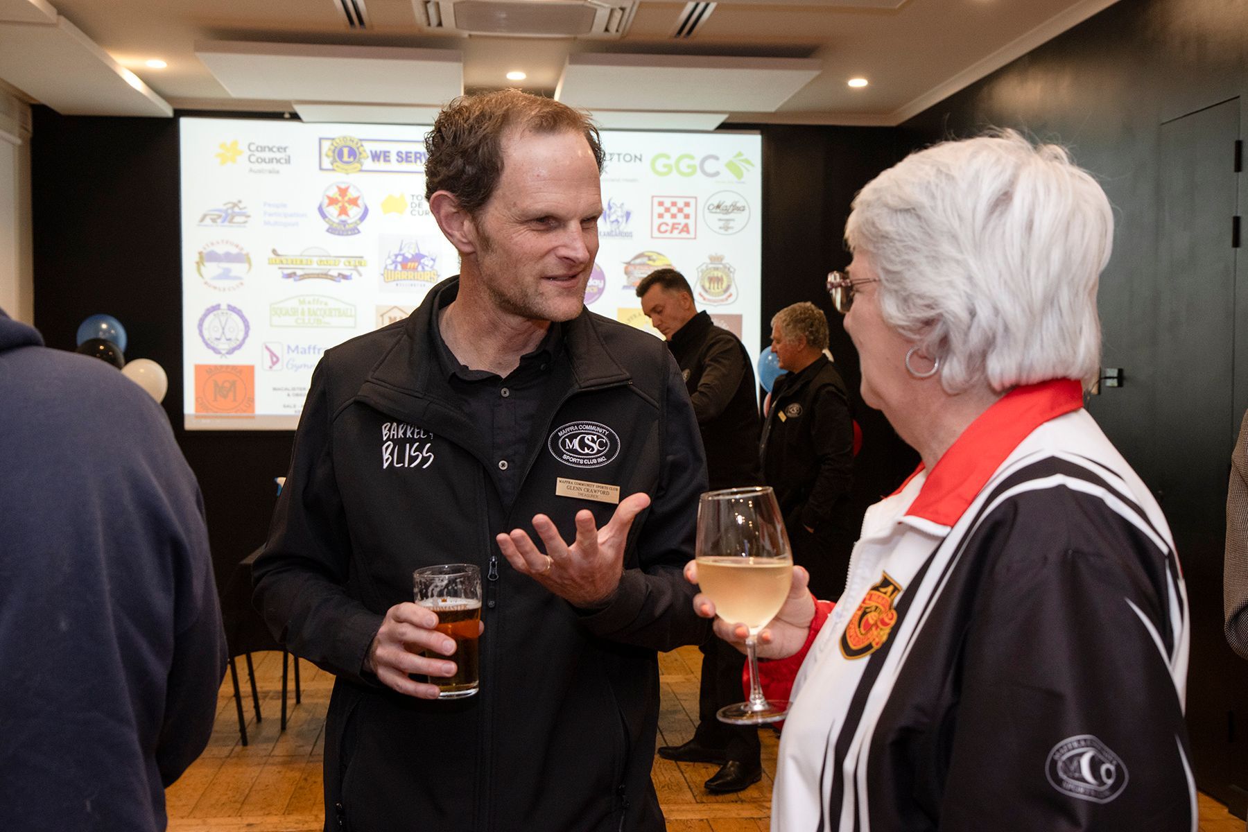Man and woman in a room talking, holding drinks. Other people in the background. Projector screen with logos.