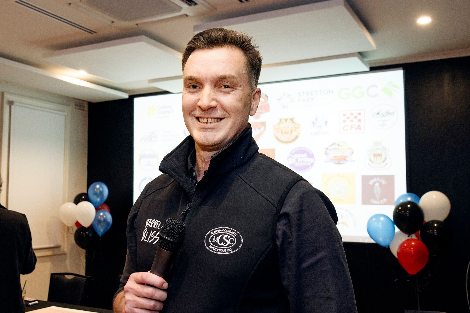 Man in black vest smiles, holding microphone; balloons in background, presentation screen.