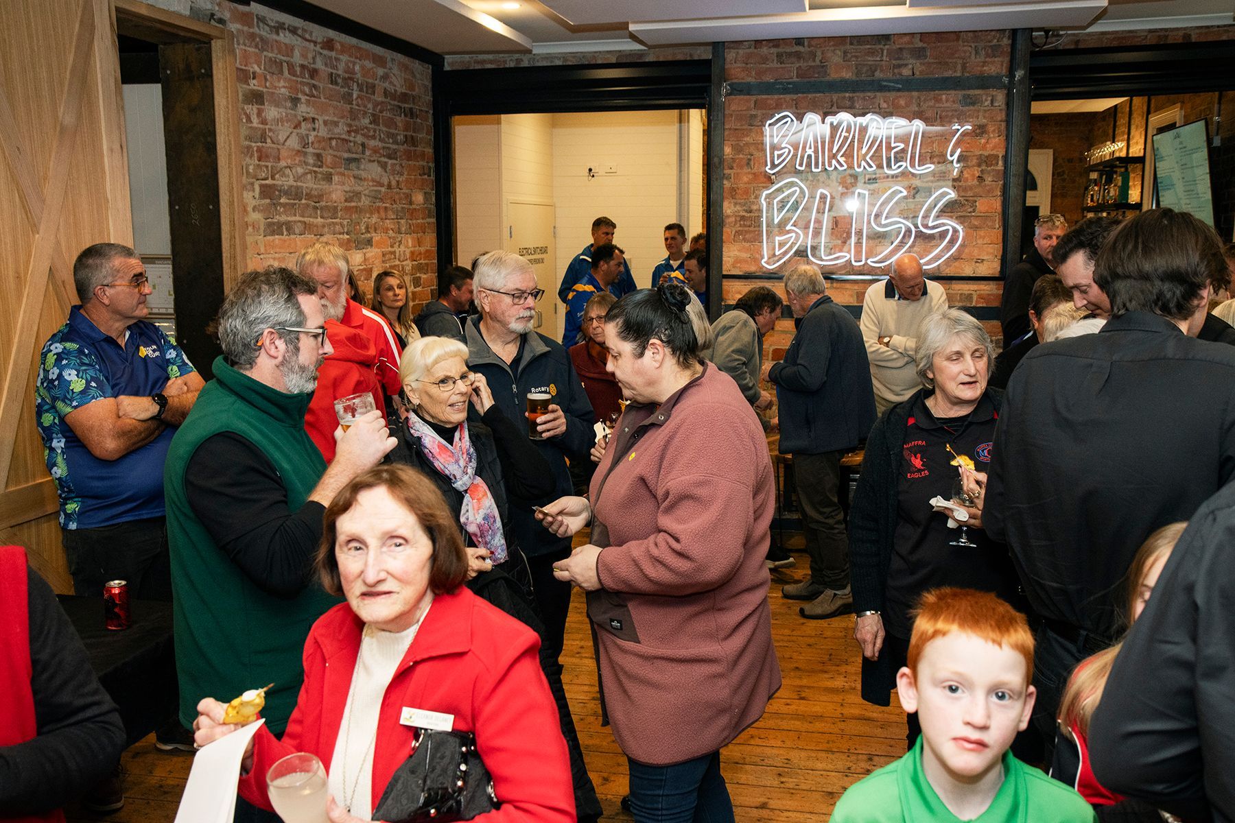 People socializing in a room, some holding drinks, brick wall background with neon sign 