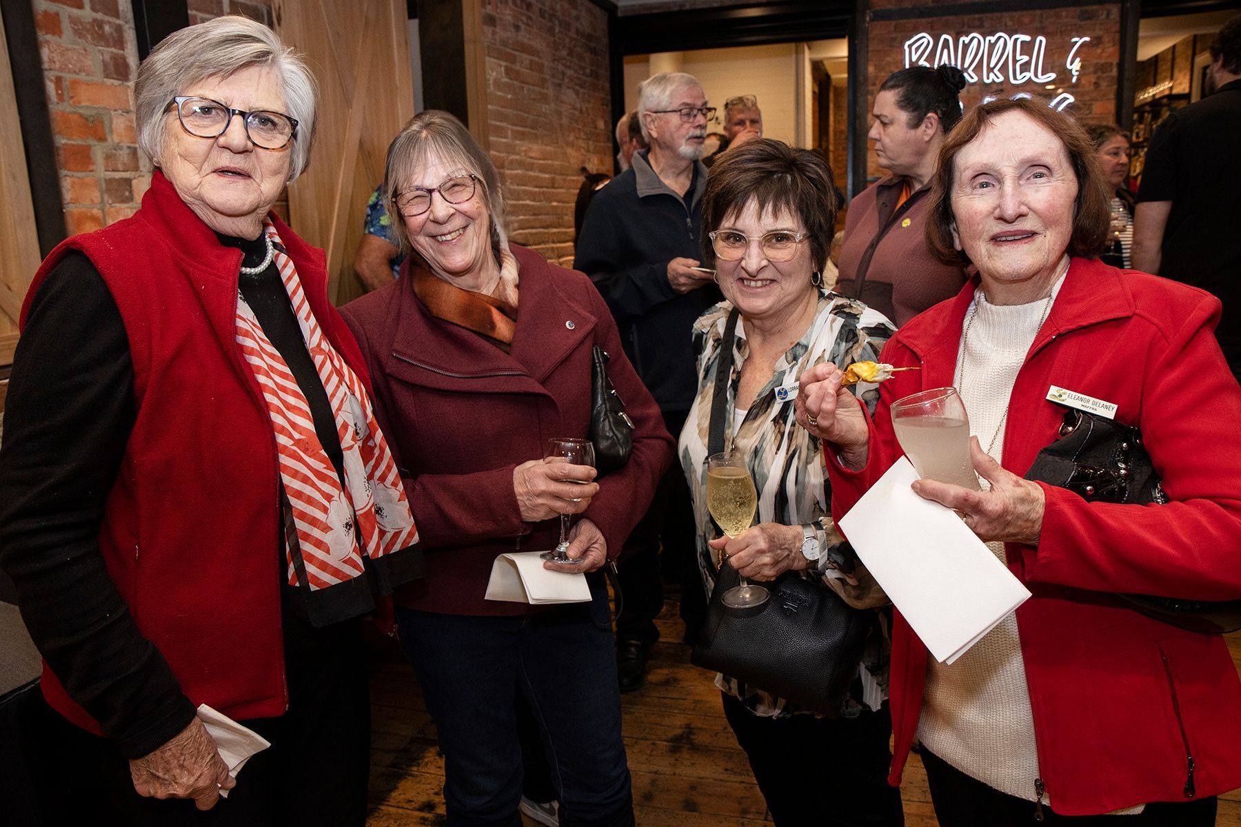 Four women smiling at an event, holding drinks, wearing red jackets, near a doorway with a sign.