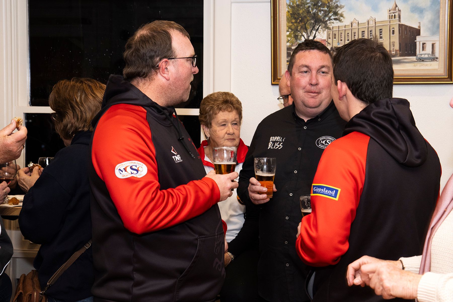 People chatting, holding drinks in a room with artwork on the wall. Men in red/black hoodies, woman with blonde hair.