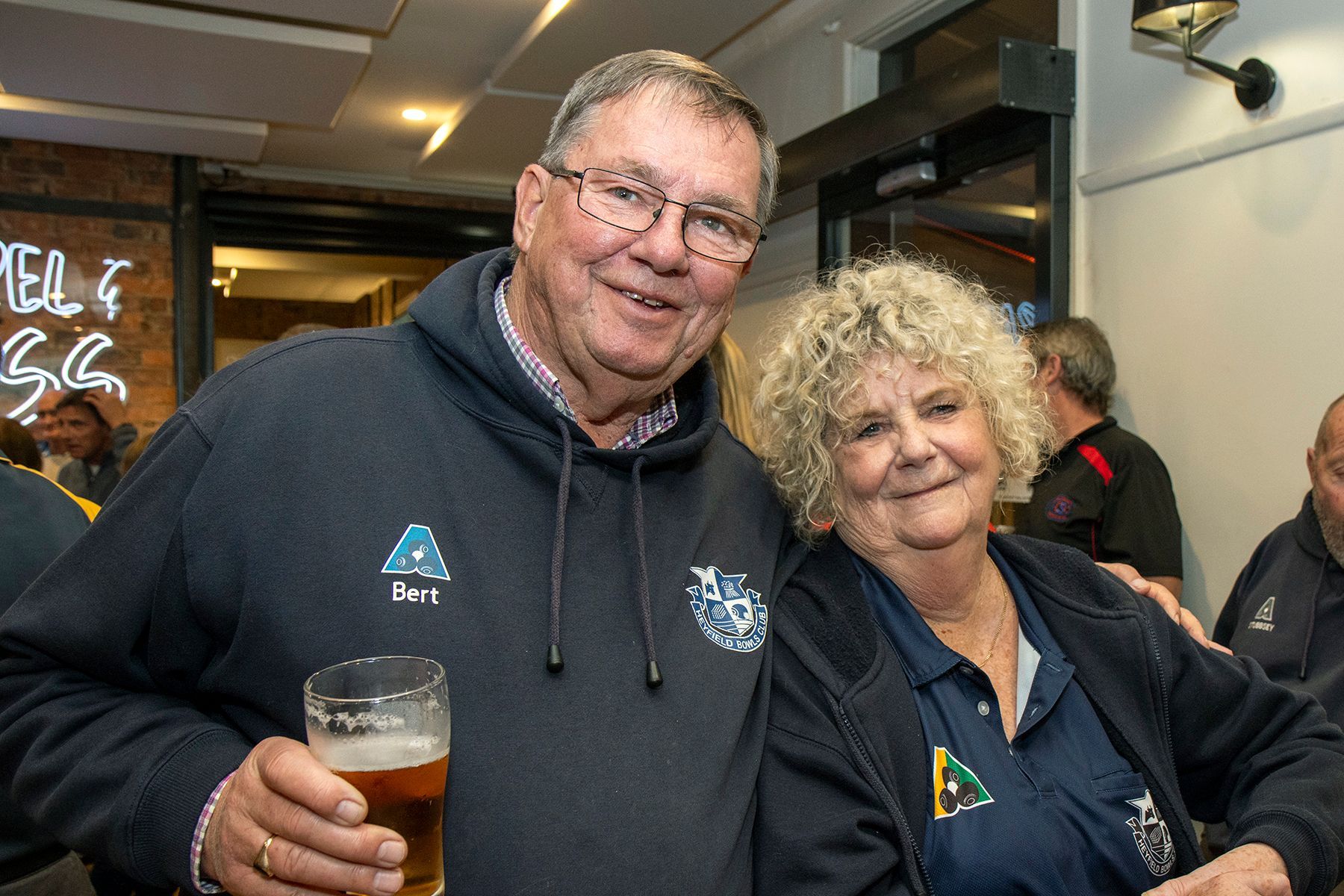 Man and woman smiling, indoors. Man holds beer, wearing a blue hoodie. Woman has curly blonde hair, wearing a navy top.