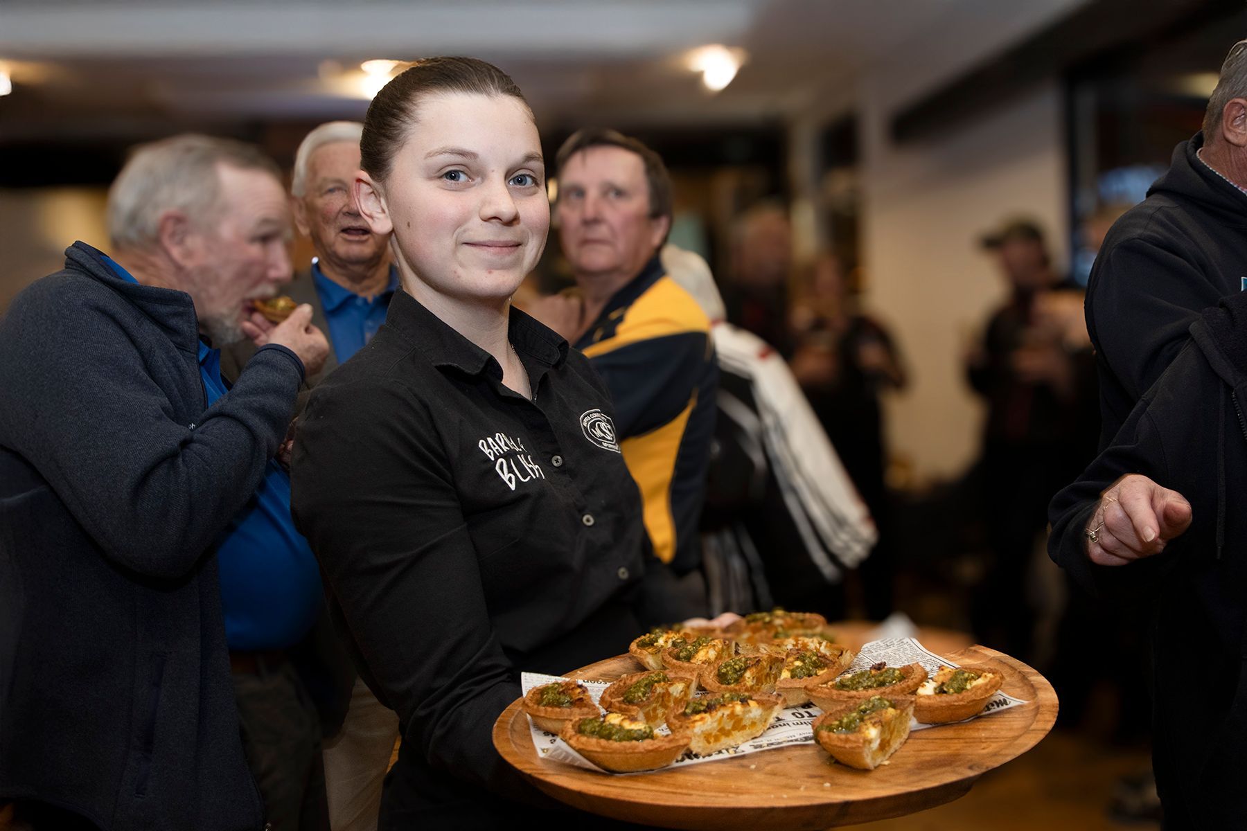 Waitress smiles, holding a tray of appetizers at an event. People gather in the background.