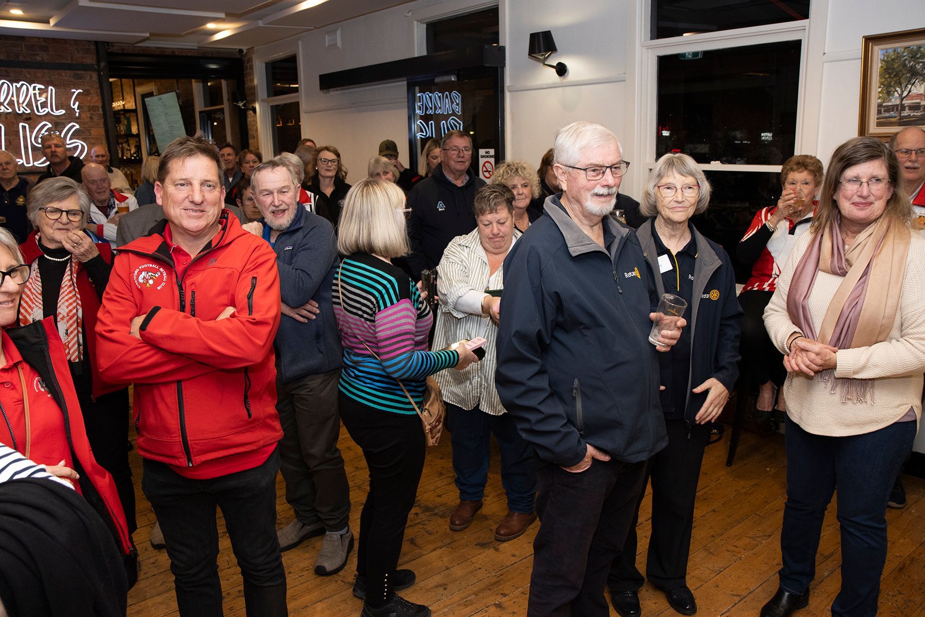 Group of people in a room, some smiling, mostly older, wearing various clothing, indoors, appears social.