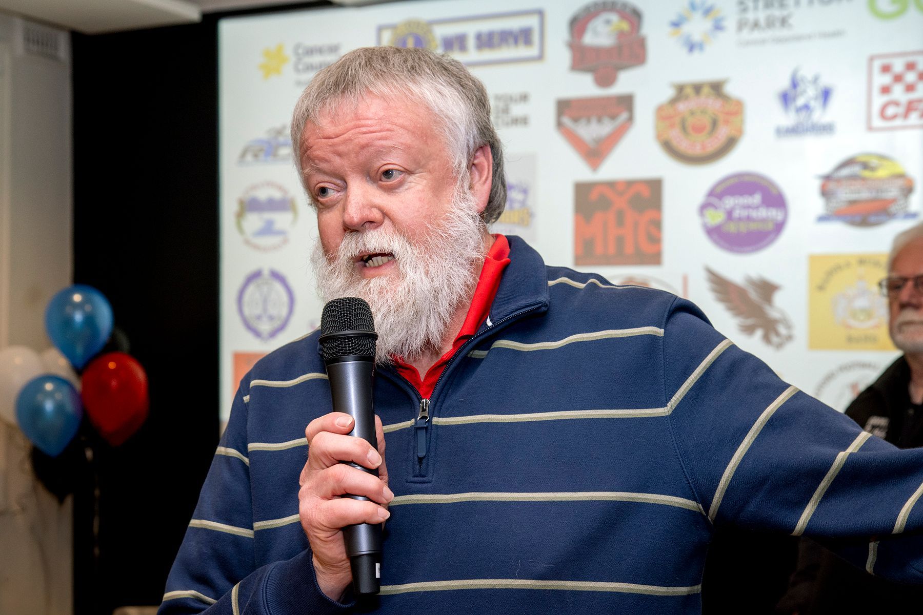 Man with white beard speaks into a microphone, blue and white striped shirt, in front of logos, indoor setting.