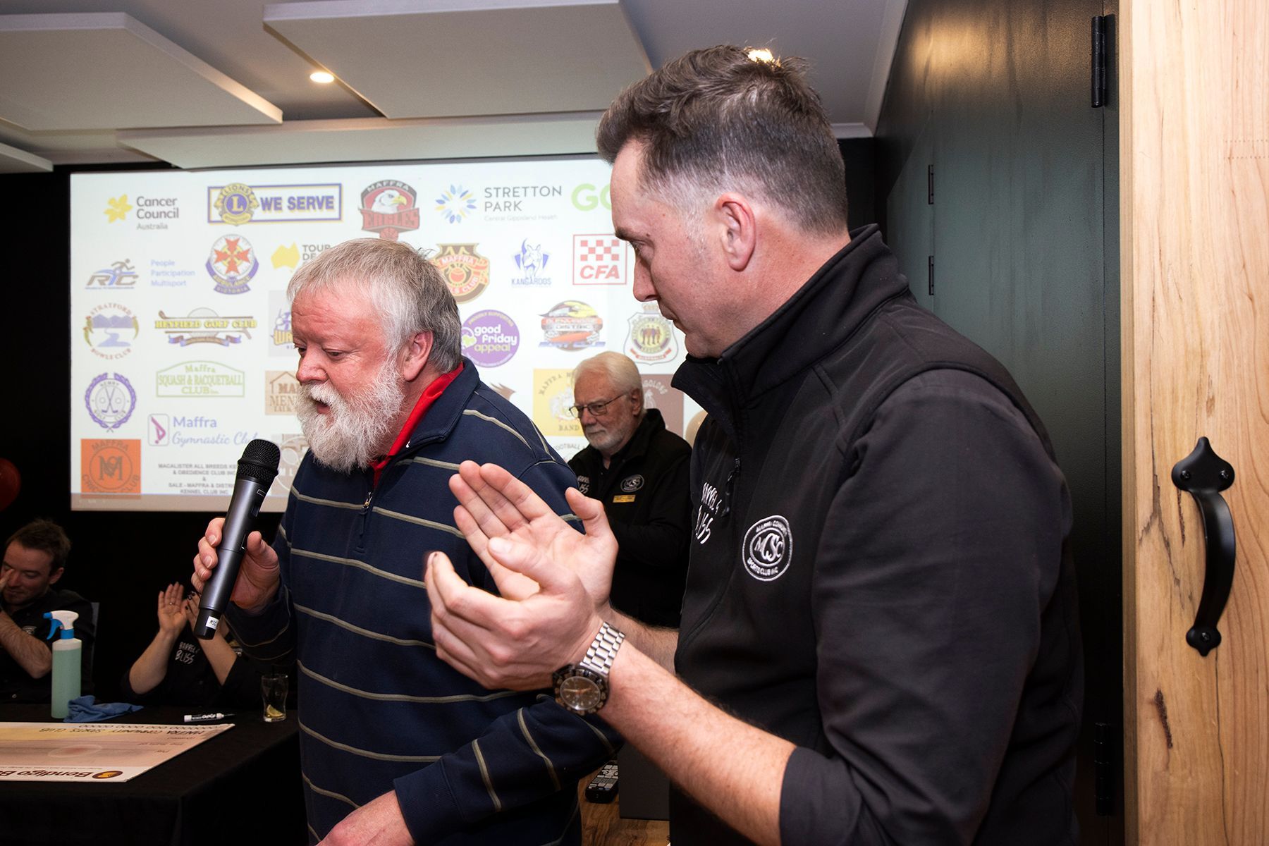 Two men at a podium, one speaking into a microphone, the other gesturing. Logos and a wood door are in the background.