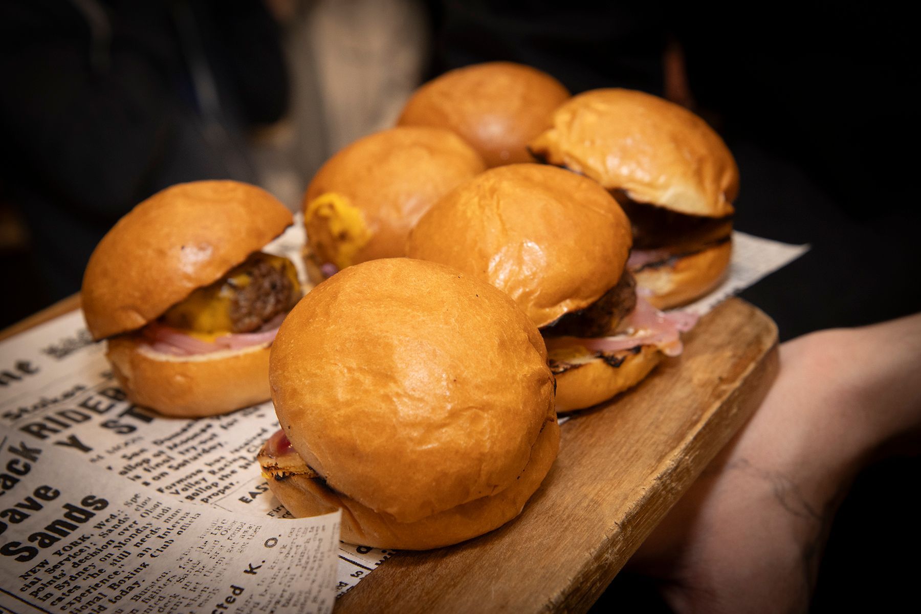 Sliders on a wooden board, with buns, patties, cheese, and fixings, held by a person.