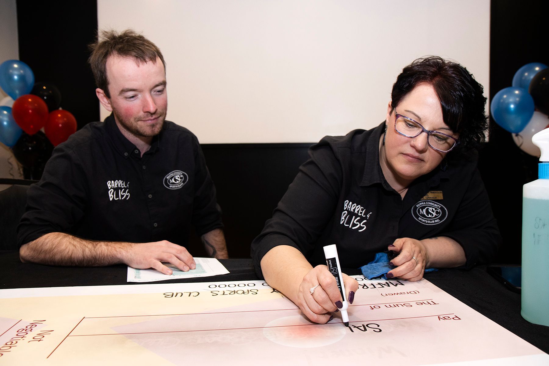 Two people sign a large check at an event; balloons in background.