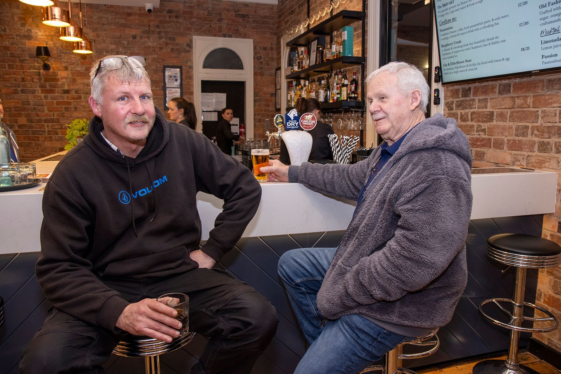 Two men at a bar, one holding a drink and smiling. They sit at a counter, near a brick wall.