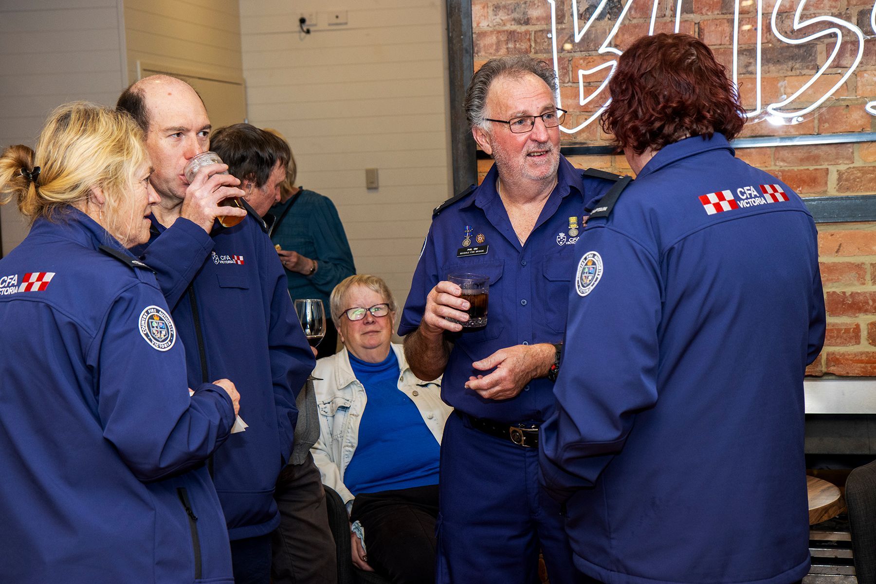 People in blue uniform socializing indoors, some holding drinks. A neon sign is visible.