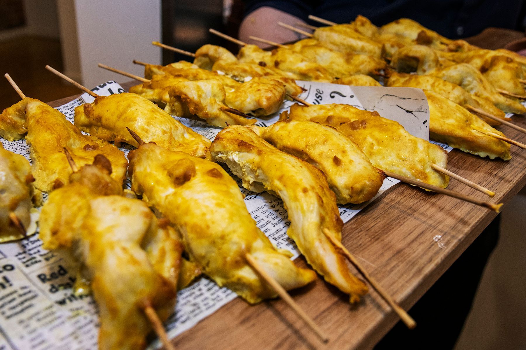 Chicken skewers on a wooden tray, golden brown, served on newspaper.