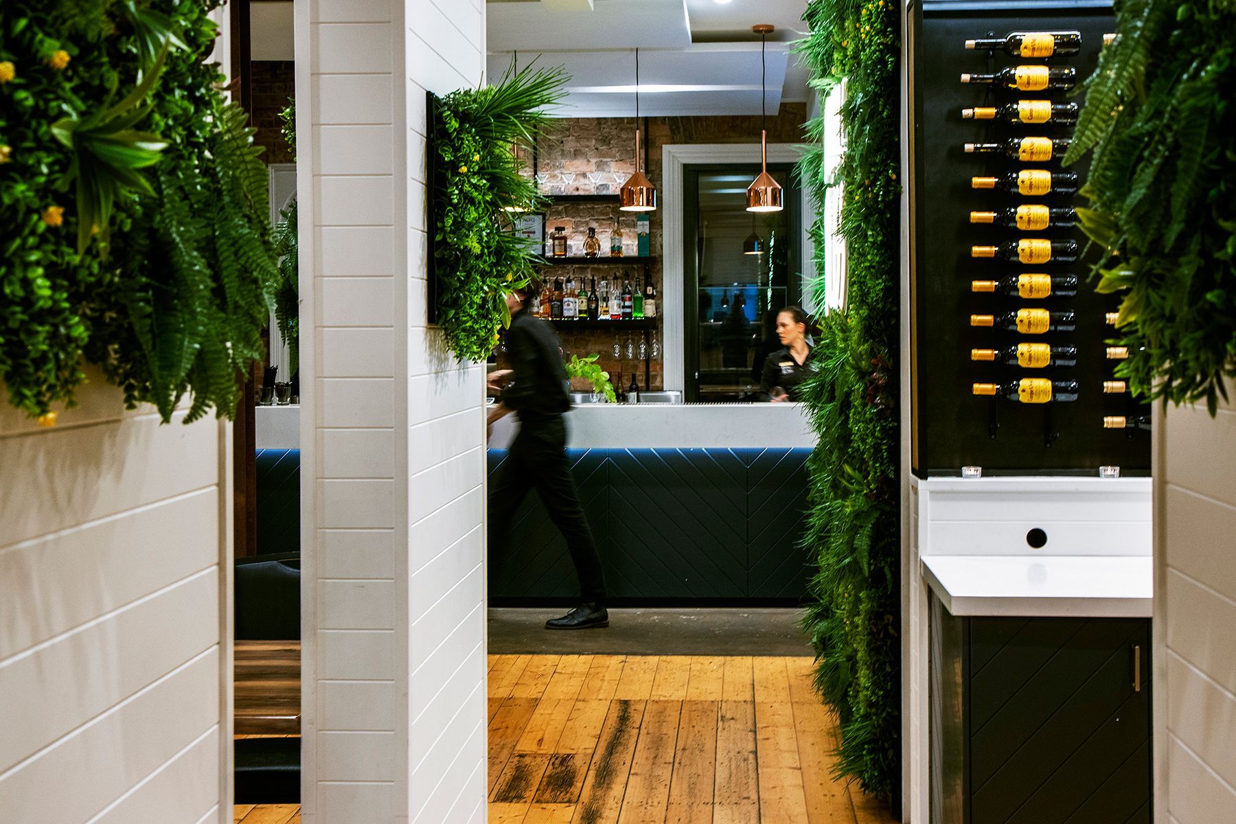 Hallway leading to a bar with greenery, a bartender, and a wine display.