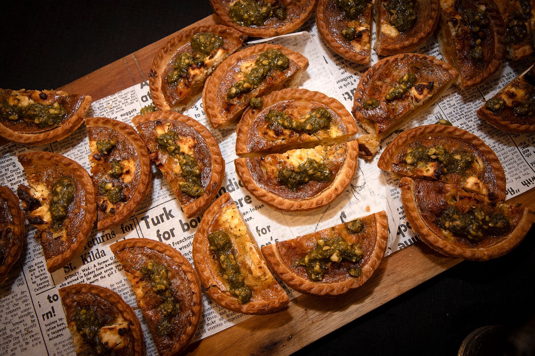 Mini quiches topped with green sauce, arranged on newspaper-lined wooden tray.