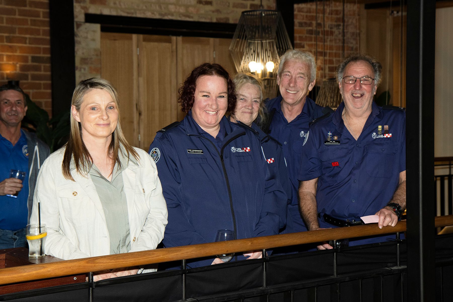 Group of people at an event, some in blue uniforms, leaning on a railing, smiling and looking at the camera.