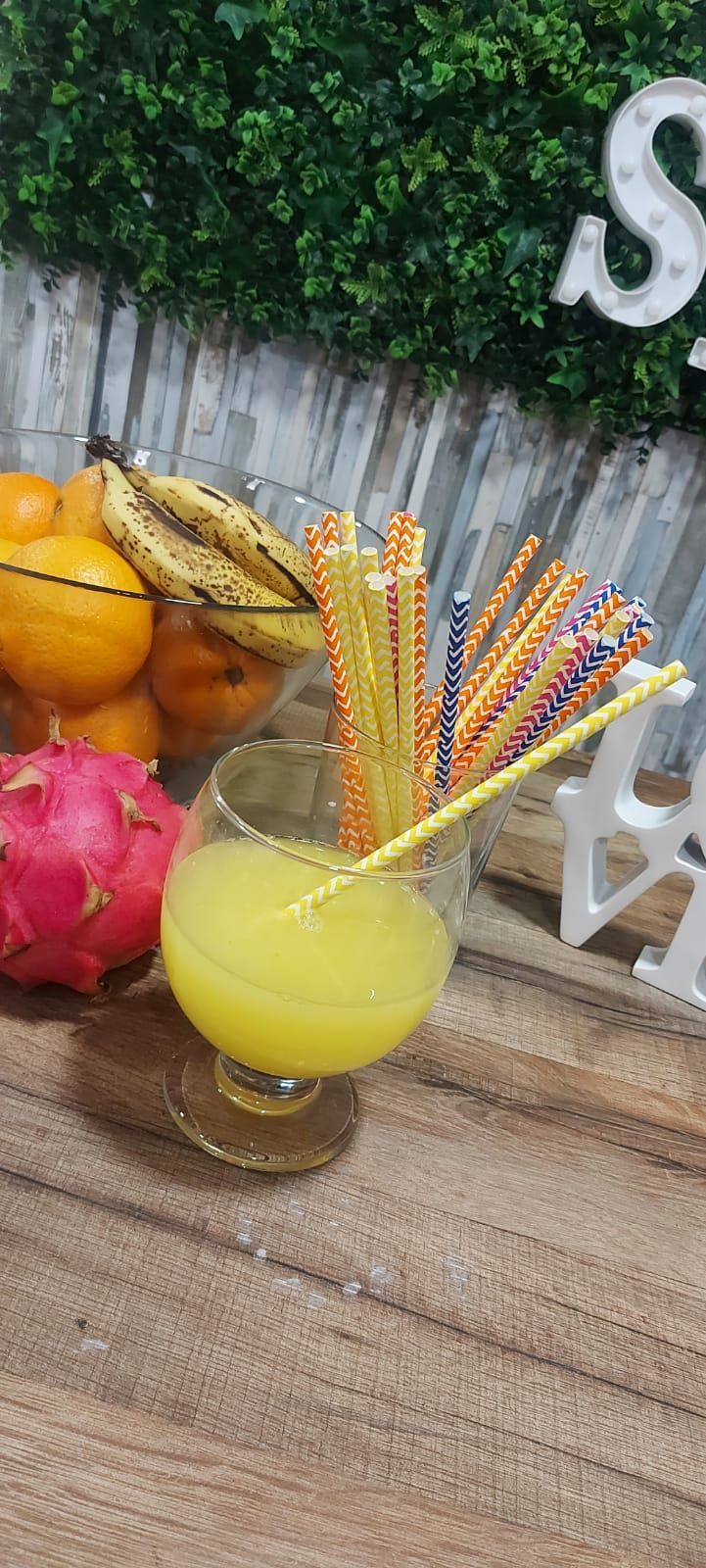 Un vaso de jugo amarillo con pajitas, fruta del dragón, naranjas, plátanos y vegetación sobre una superficie de madera.